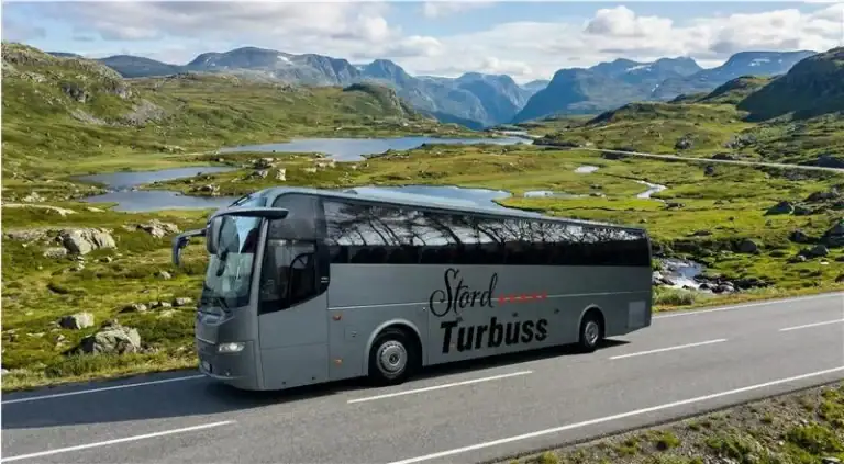 En moderne turistbuss fra Stord Turbuss kjører gjennom naturskjønt landskap med fjell og innsjøer, perfekt for gruppereiser og fjellturer.